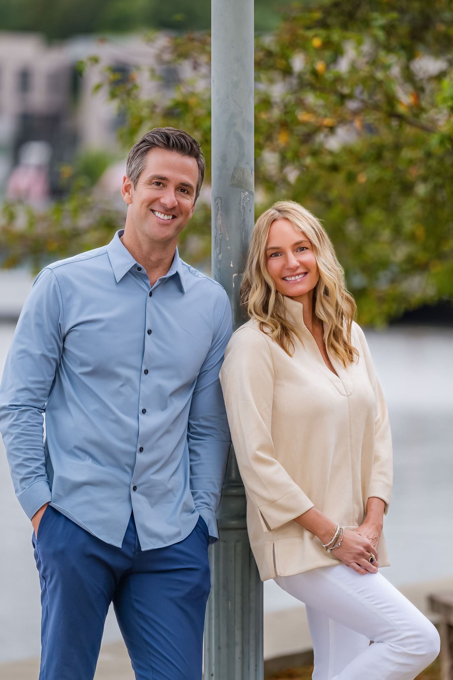 A smiling man and woman pose casually by a lamppost outdoors near a waterfront on a sunny day.