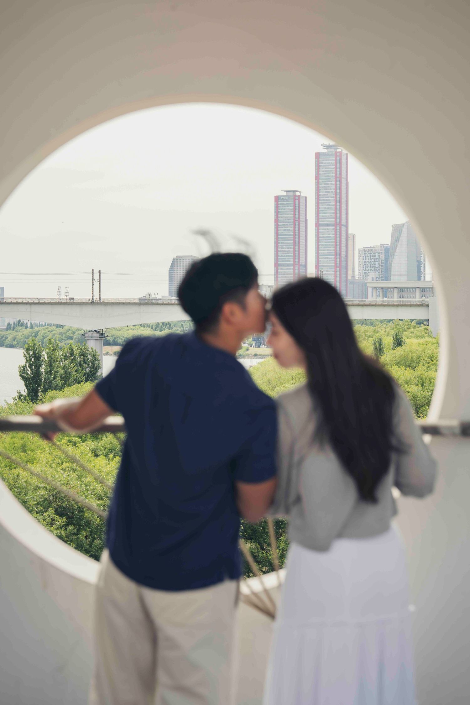 Couple sharing a romantic moment framed by a circular window overlooking a city skyline and lush greenery.