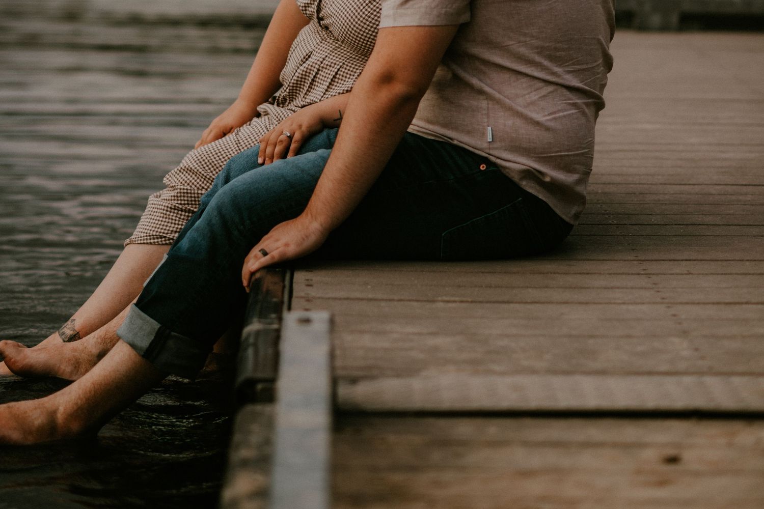 Couple sitting closely together on a wooden dock, legs intertwined, sharing an intimate moment by the water.