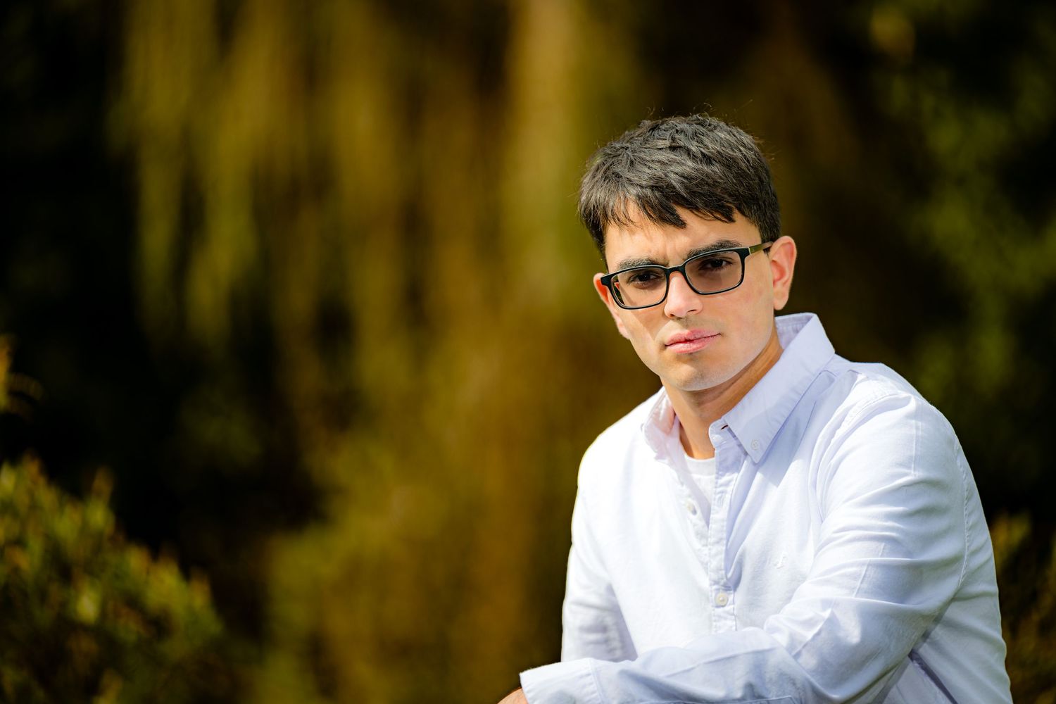 Young man wearing sunglasses and white shirt sitting outdoors with green foliage background.