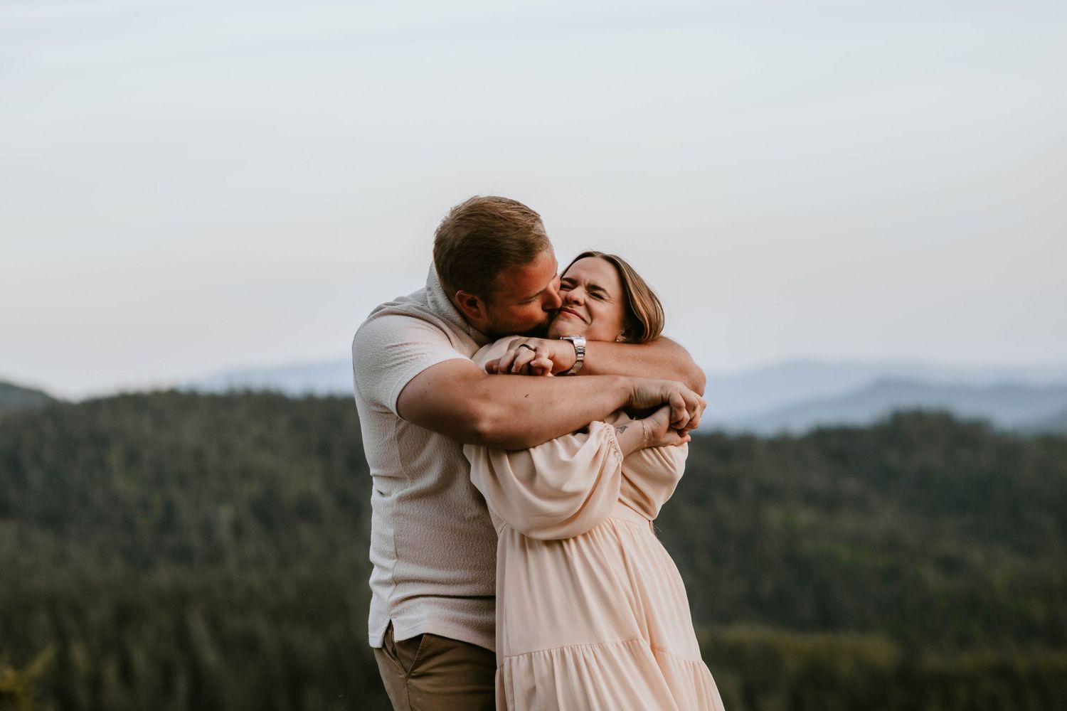 Couple embracing and laughing on a mountain overlook with forested hills in the background at dusk.