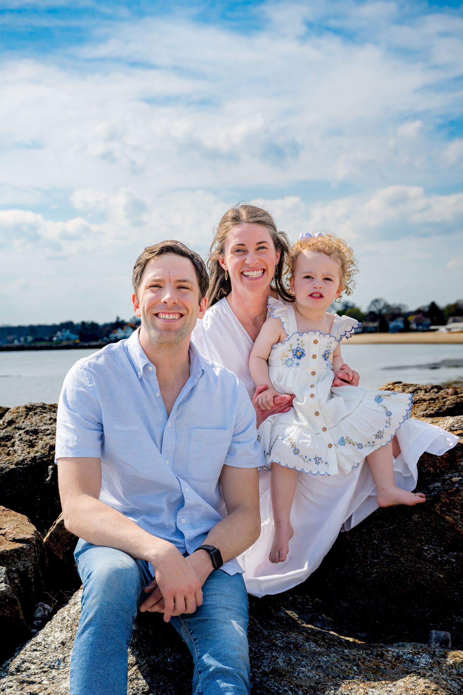 A smiling family of three poses on rocky beach, parents in white and blue with toddler girl in floral dress.
