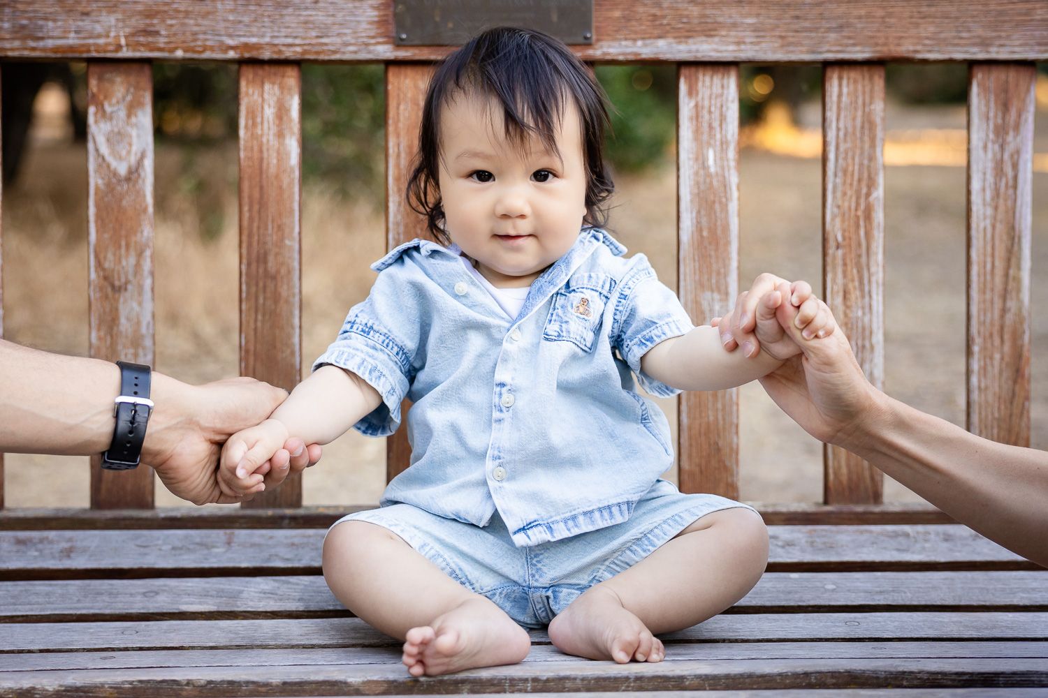 Cute six-month baby sitting cross-legged on wooden bench holding hands with parents at his milestone photography session in Palo Alto