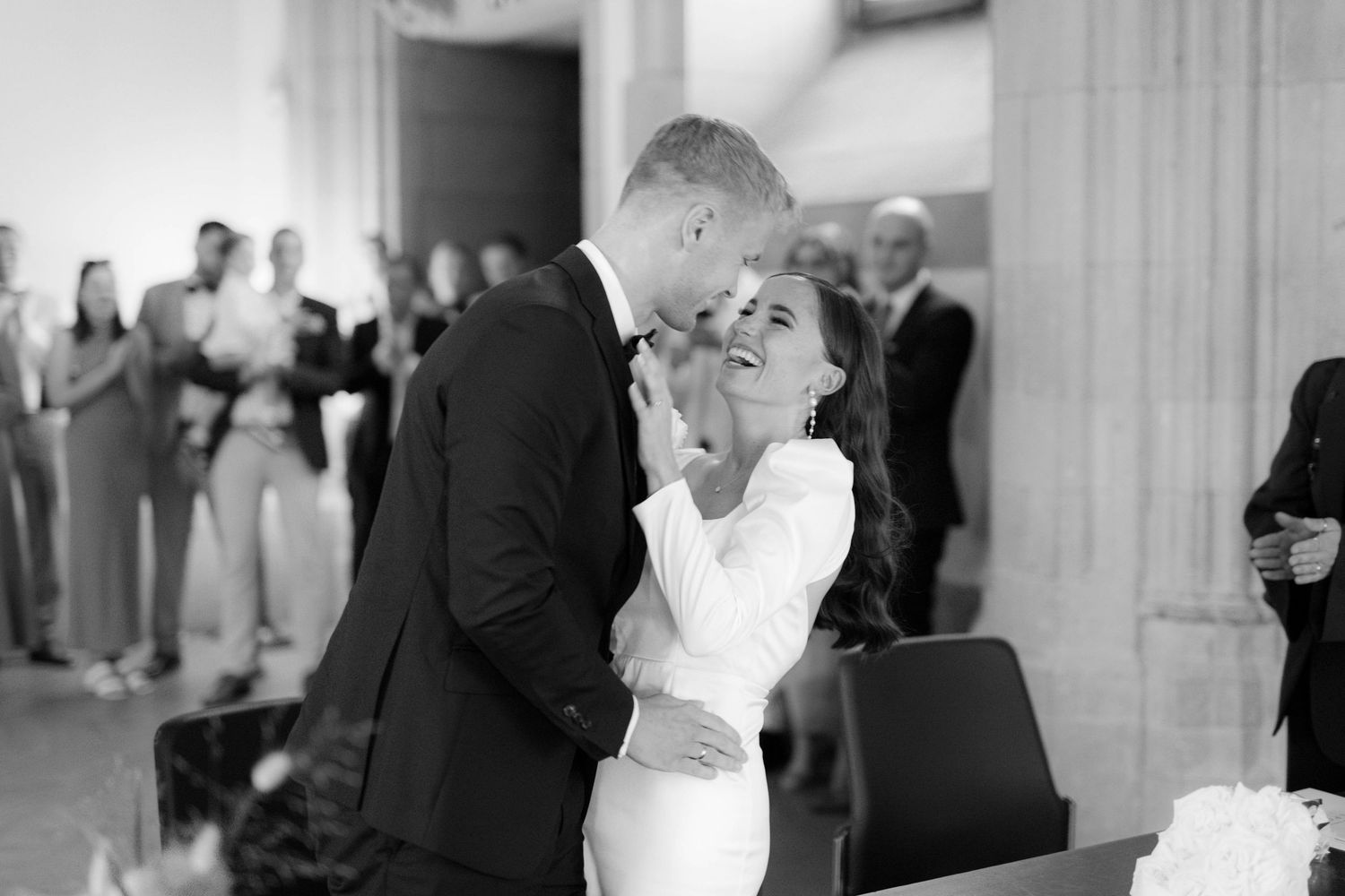 Black and white photo of a bride and groom sharing a tender moment during their wedding ceremony with guests in background.