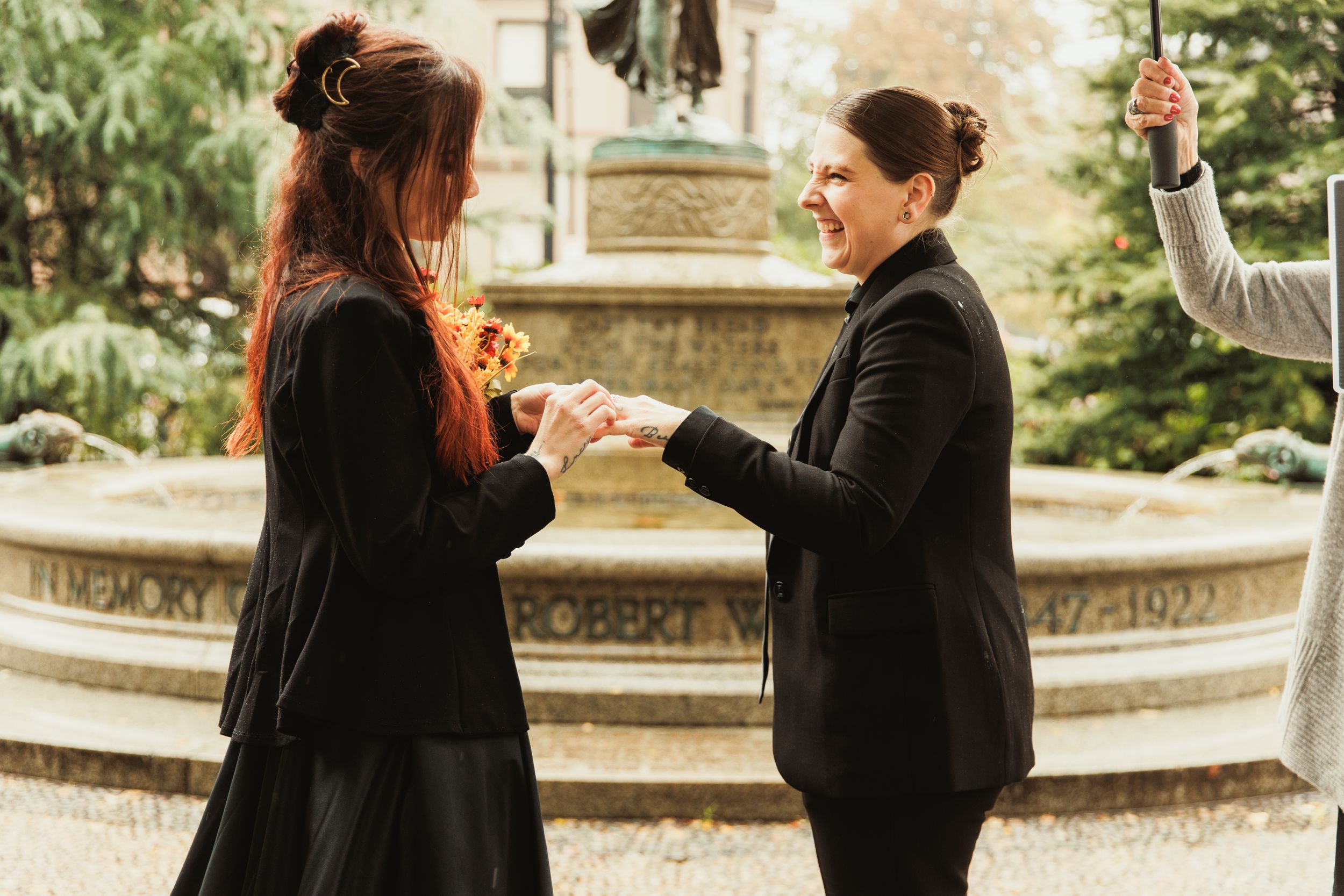 Two women in black suits exchange rings by a fountain in a romantic outdoor same-sex wedding ceremony.