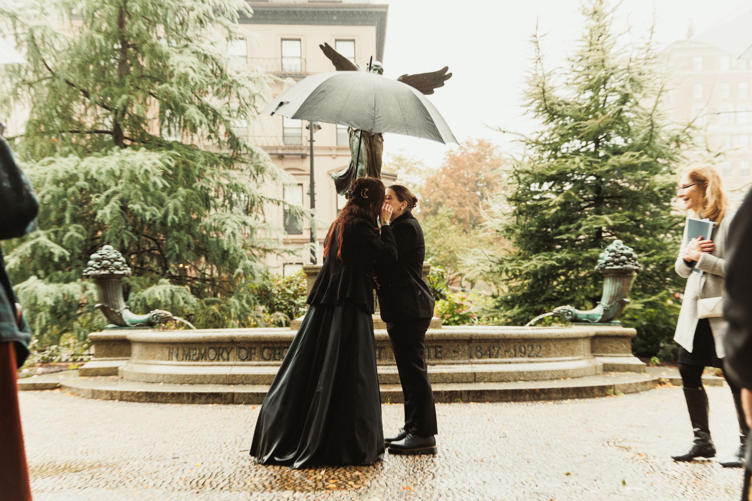 Couple kissing under umbrella in rain by fountain, one wearing black gown, in lush garden setting.