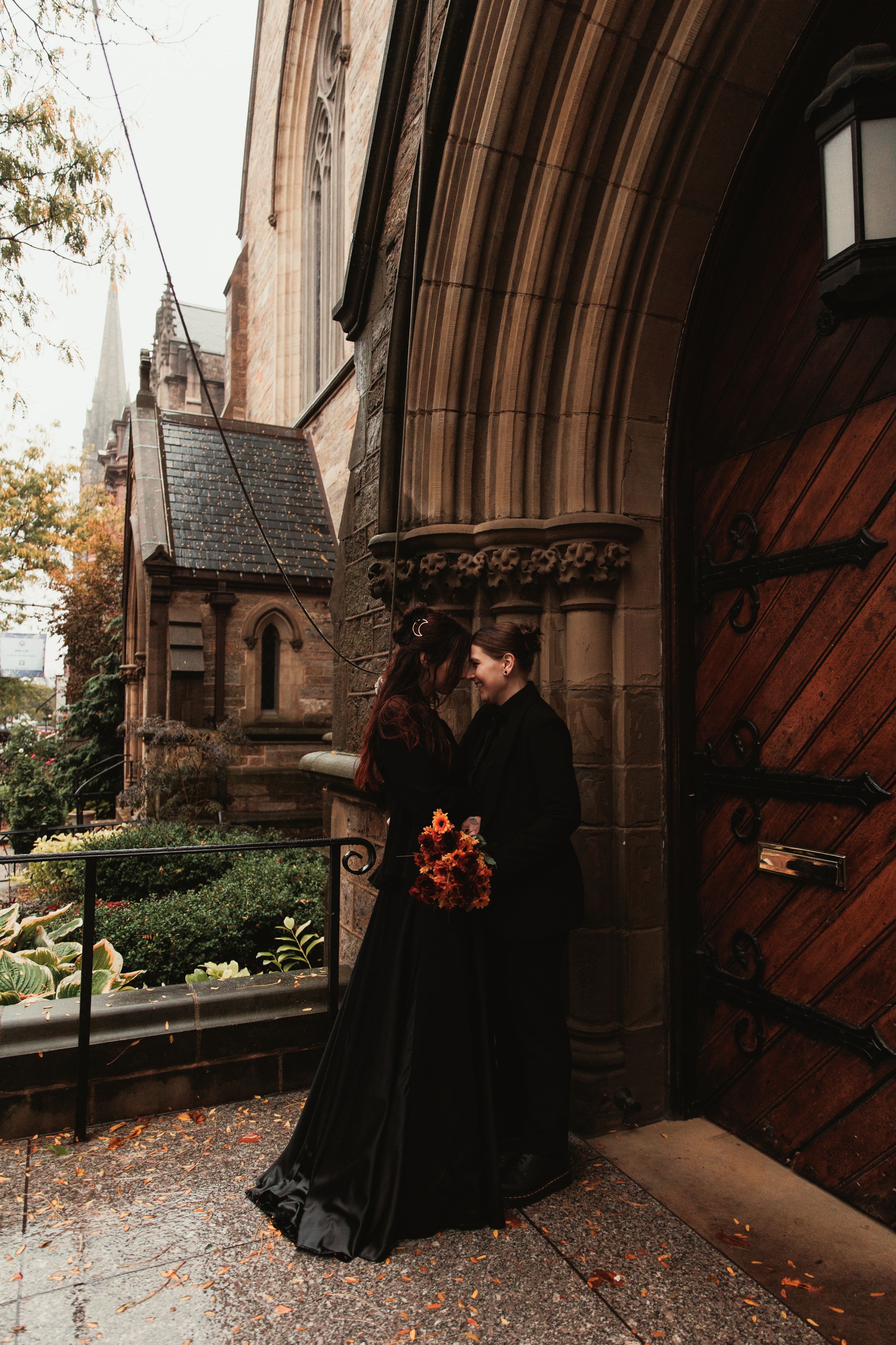 Couple in black wedding attire embracing at gothic church entrance with autumn foliage and ornate wooden doors.
