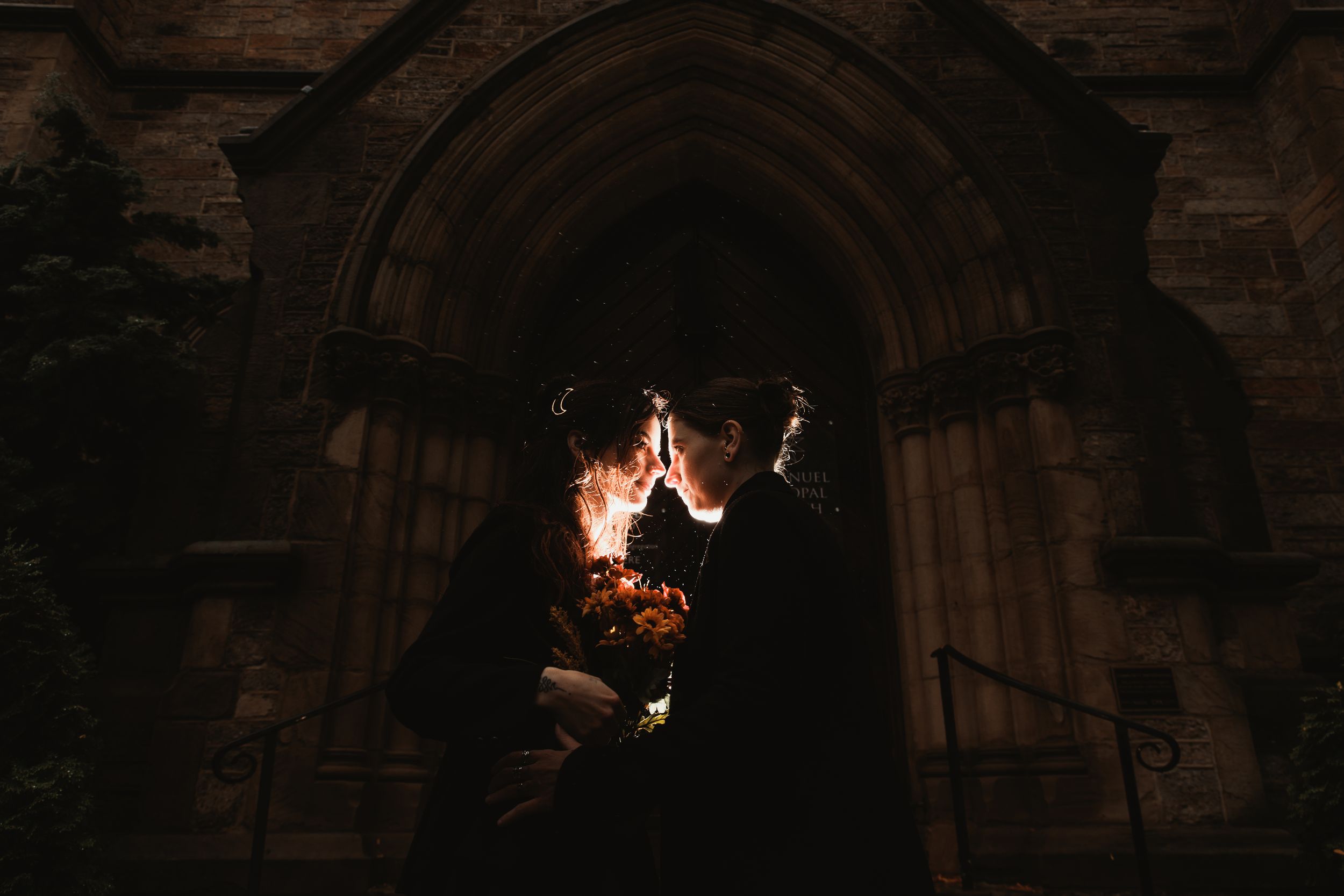 Two brides in black share a romantic kiss before a gothic church archway, holding a vibrant red bouquet.