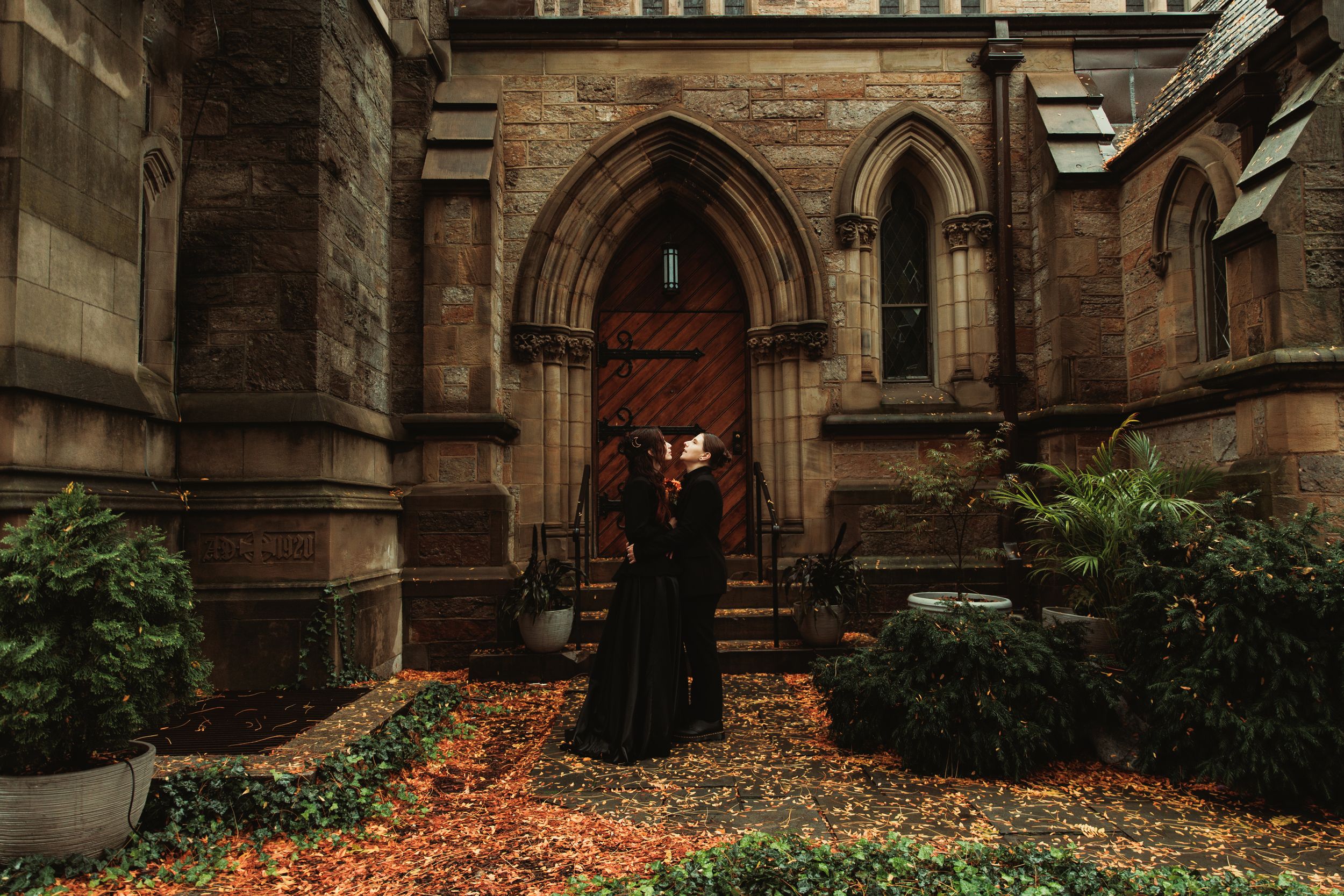 A person in a black gown stands before a Gothic stone church entrance surrounded by autumn leaves.