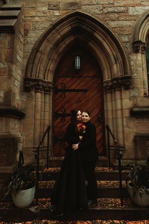 Couple in matching black wedding attire embracing before Gothic stone church doorway with ornate arch.