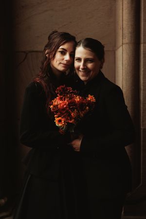 Two brides in matching black coats smile together, holding a stunning bouquet of orange and red flowers.