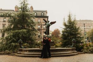 Couple in black wedding attire embracing near angel fountain surrounded by evergreen trees in urban park setting.