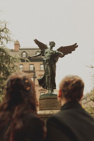 Two people admire a bronze winged angel statue on a pedestal in a park, with a historic building in the background.