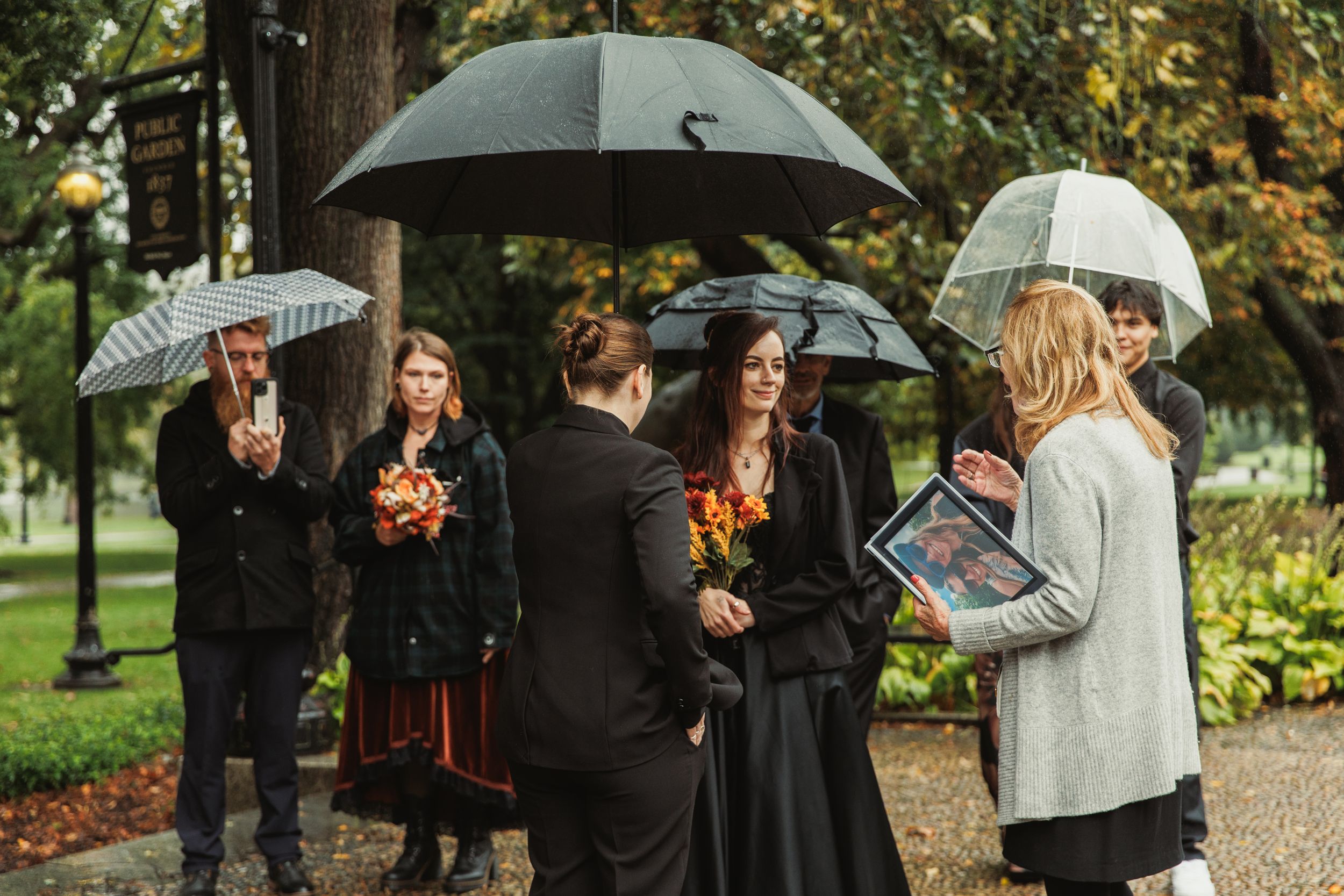 Group of people holding umbrellas in the rain at an outdoor ceremony, dressed in dark formal attire.