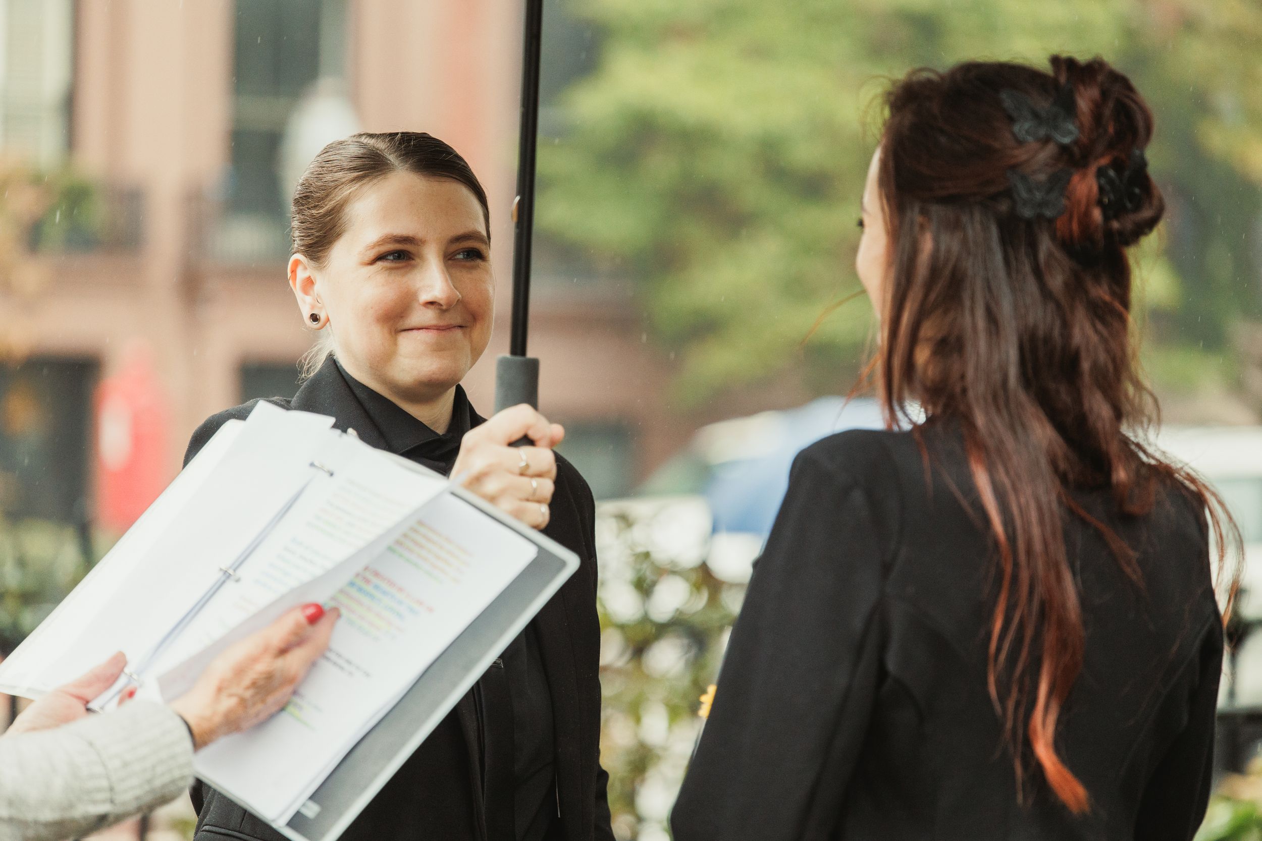 Two professional women in business attire discussing documents on a clipboard outdoors.