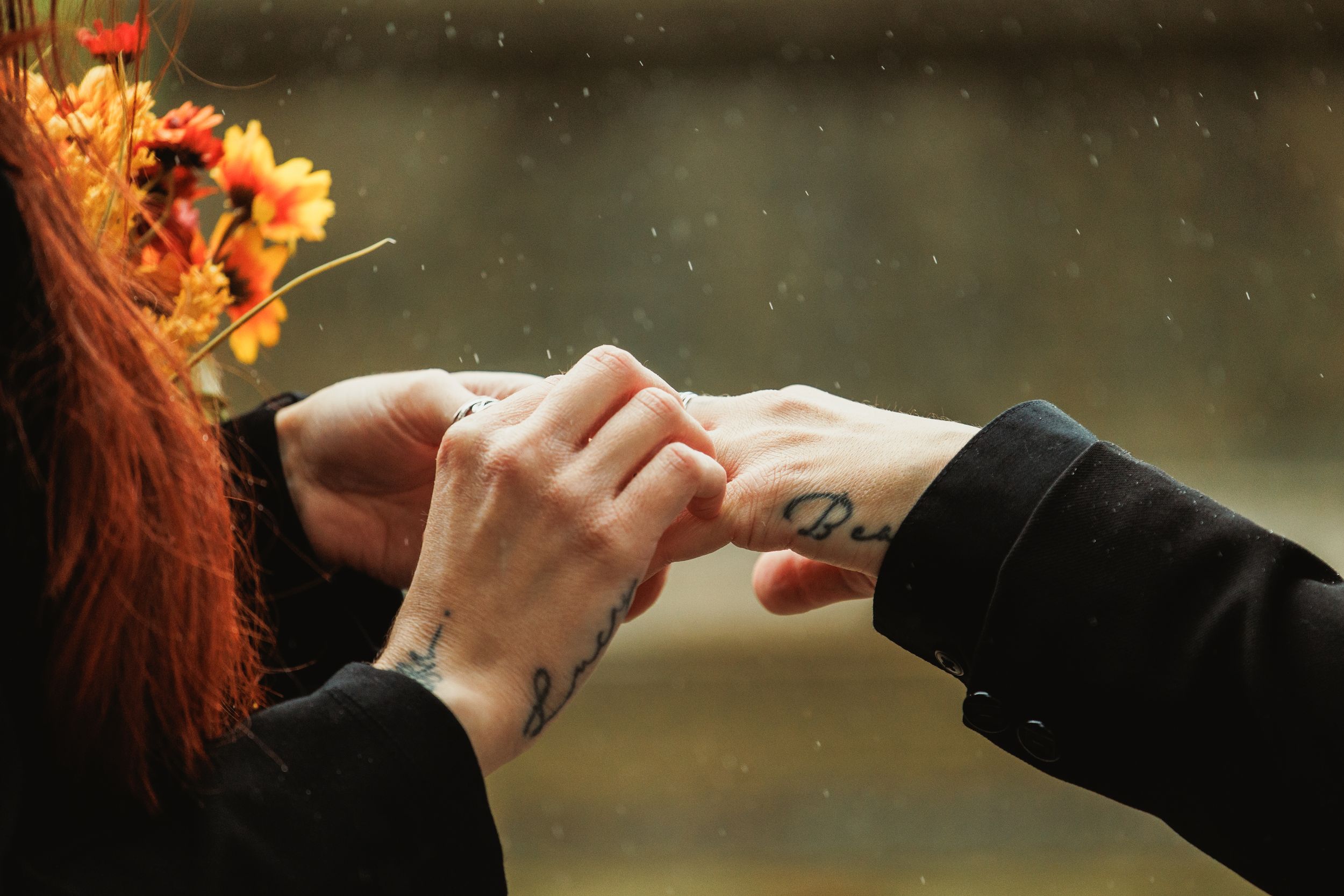 Two people with tattoos hold hands tenderly in the rain, one wearing a floral crown of autumn flowers.
