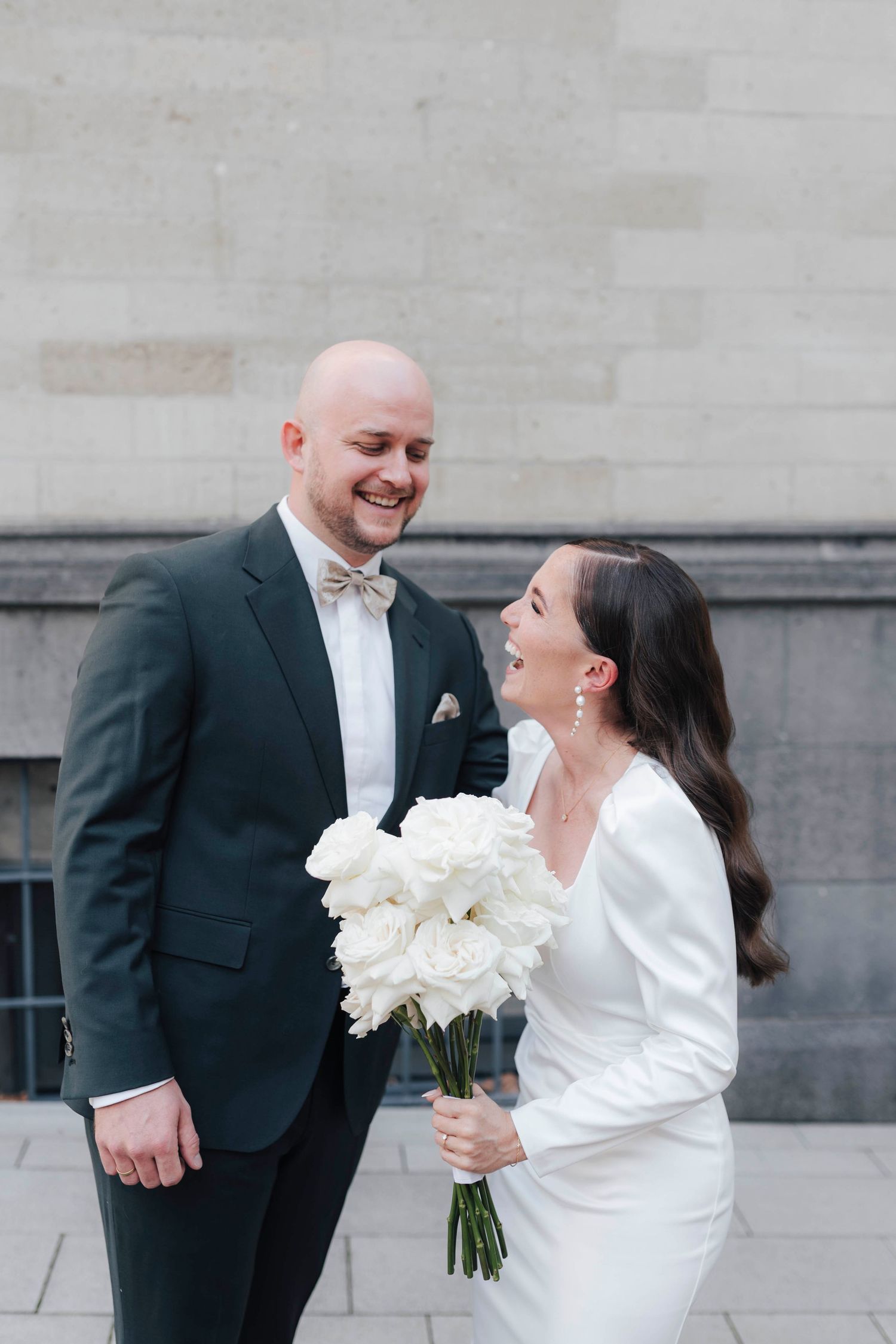A smiling bride in a white dress holds white roses while laughing with her bald groom in a dark suit and bow tie.