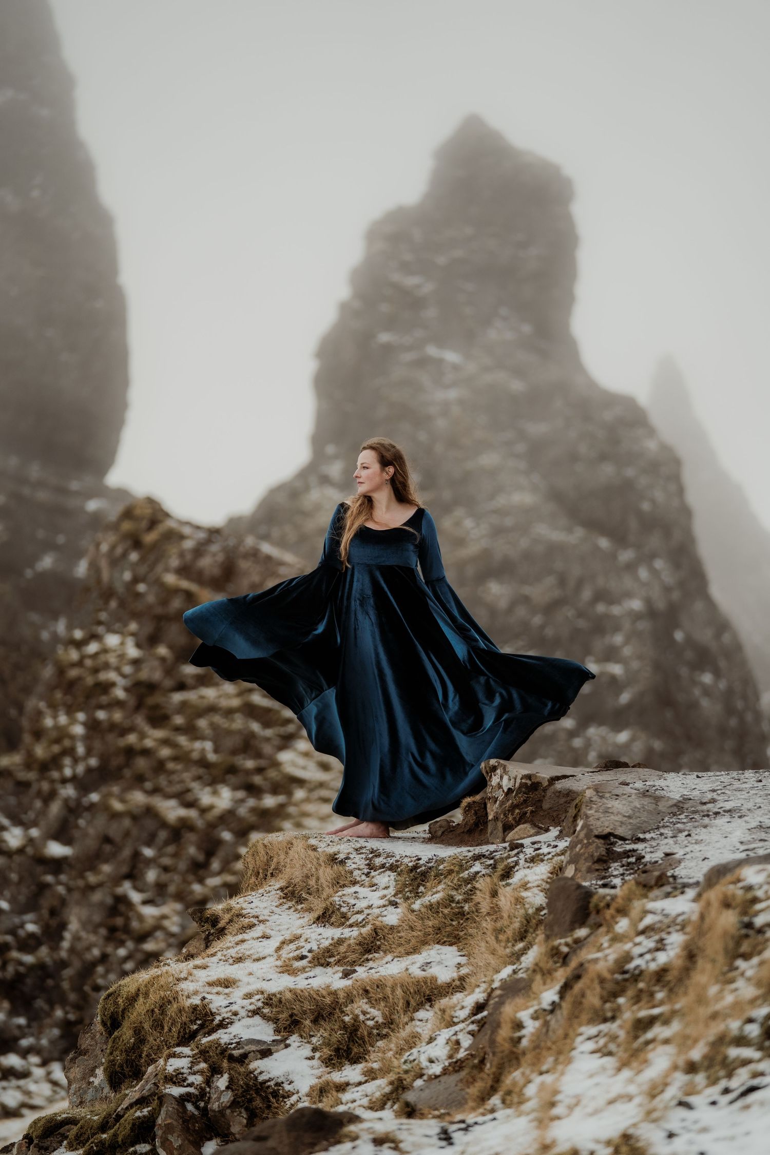 Woman in flowing dark dress walking barefoot on a black sand beach with dramatic waves and rocky cliffs.