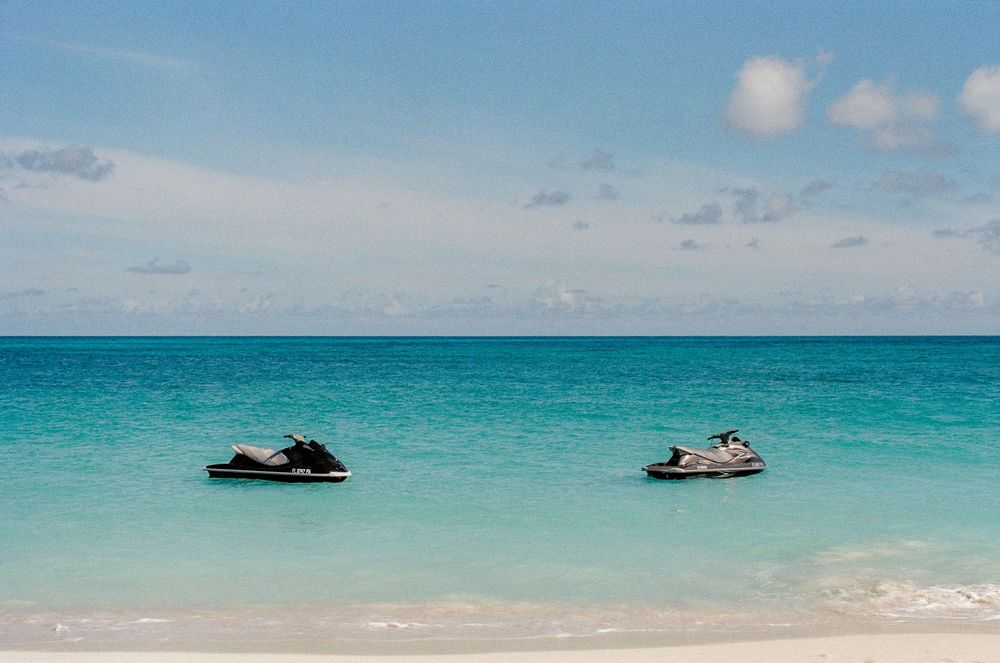 Two jet skis floating on crystal-clear turquoise water near a white sandy tropical beach under a partly cloudy sky.