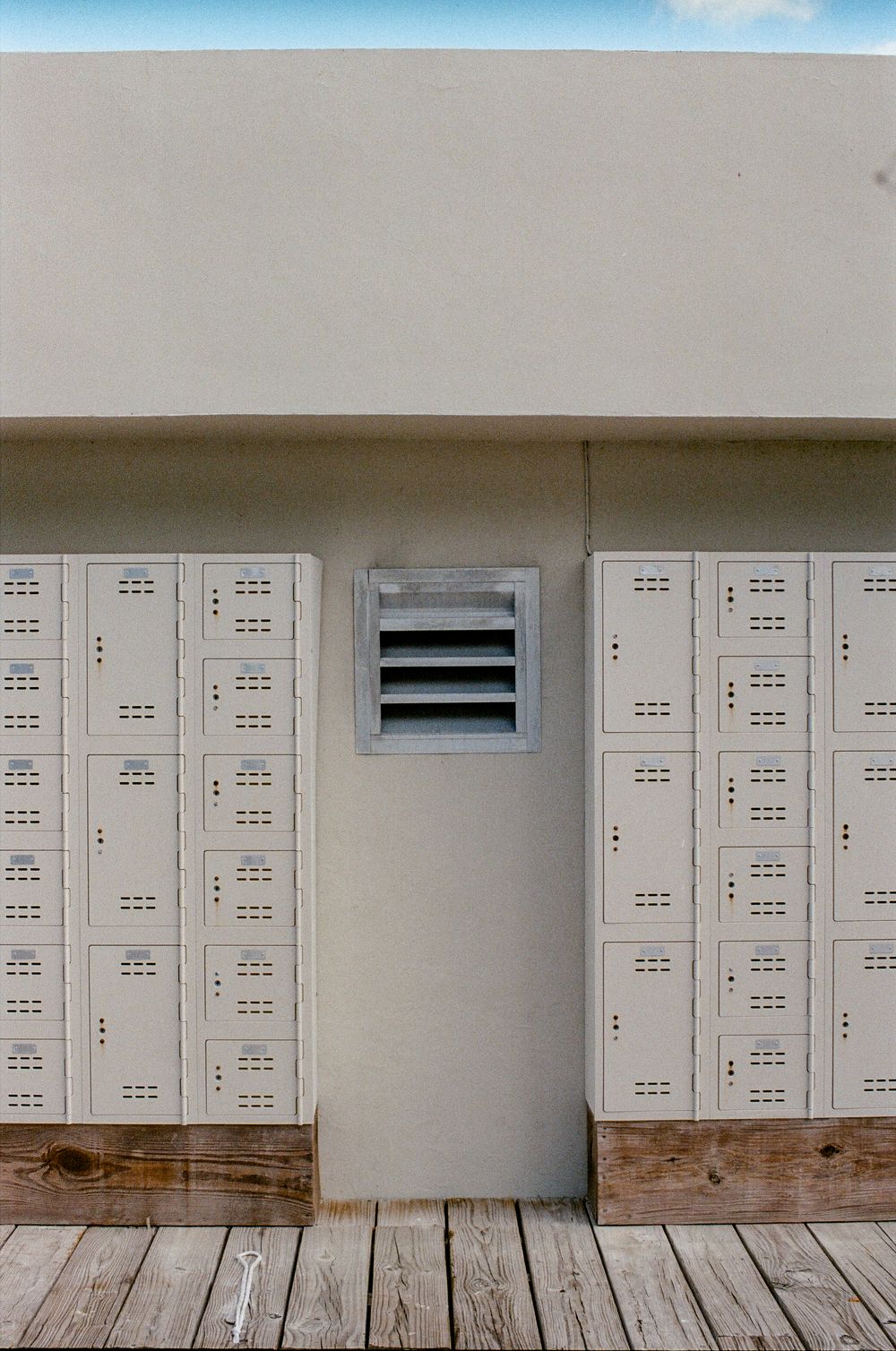 Rows of gray metal lockers on a wooden deck against a beige wall with a ventilation vent.