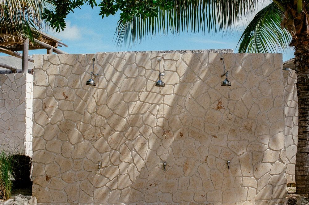 Outdoor stone wall shower with multiple shower heads near a tropical palm tree under a blue sky.