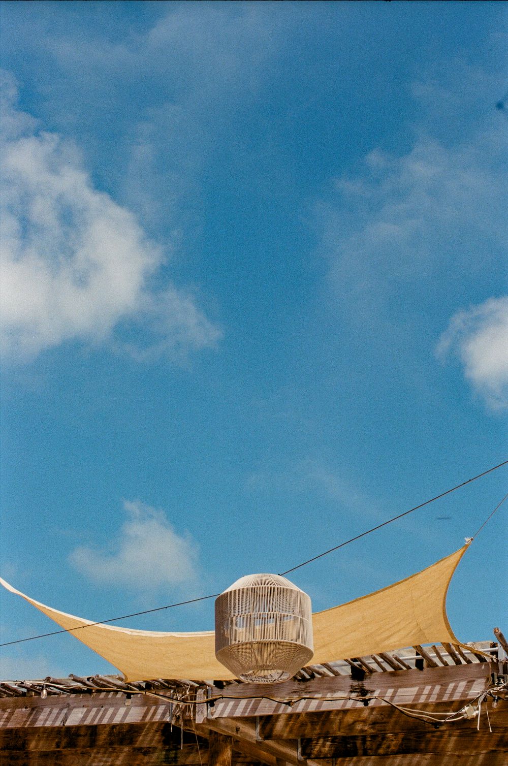 A yellow shade sail and wicker lantern against a bright blue sky with white clouds.