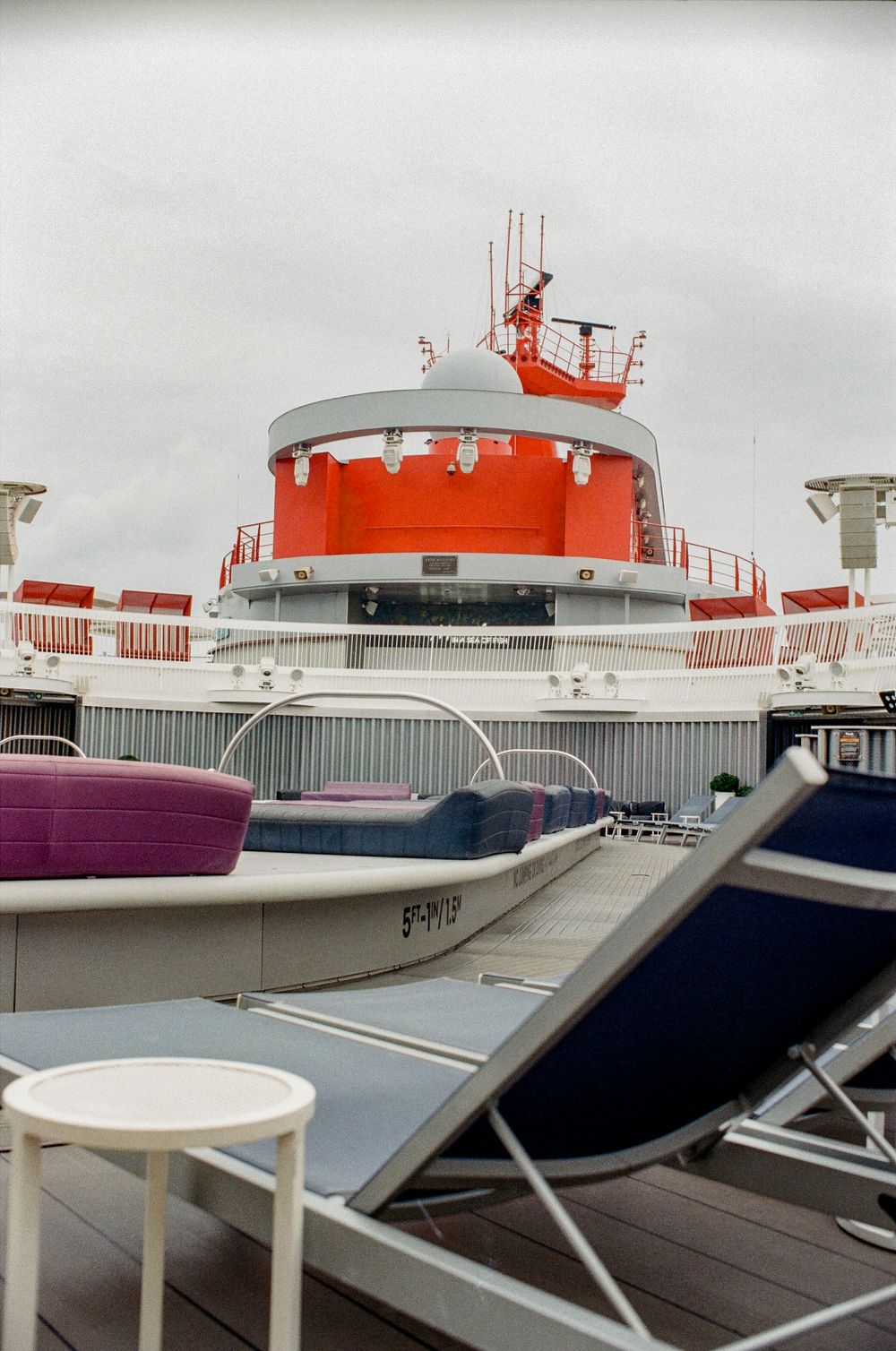 Cruise ship deck with lounge chairs and a distinctive red and white funnel tower against an overcast sky.