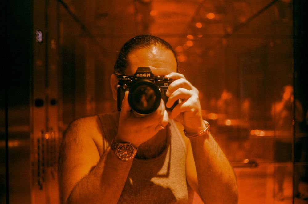 Person taking a mirror selfie with a black Pentax film camera wearing a bracelet in warm orange-tinted light.