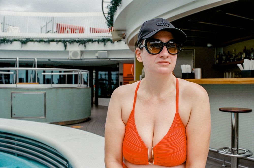Woman in orange bikini and black cap with sunglasses relaxing on a cruise ship pool deck.