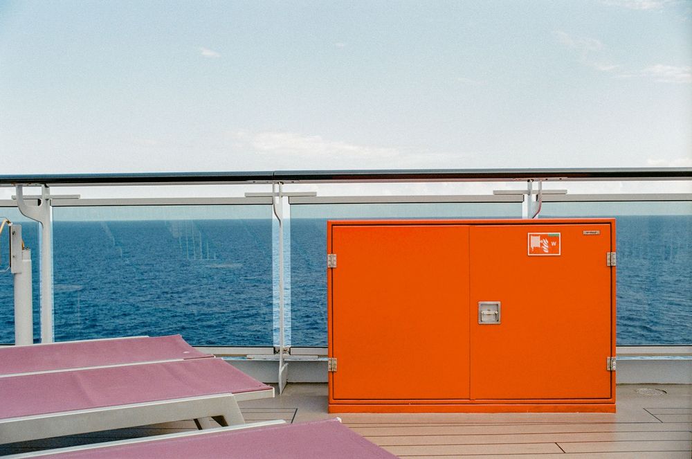 Orange storage cabinet on a cruise ship deck with ocean views, glass railing, and pink lounge chairs nearby.