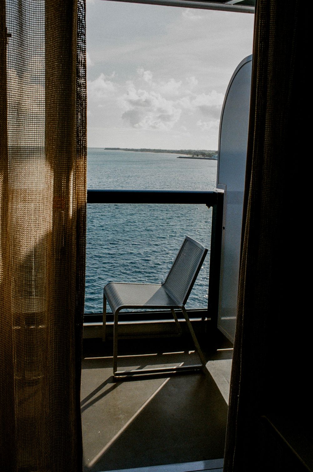 A chair on a small balcony overlooks a calm blue sea, viewed through an open door with sheer curtains.