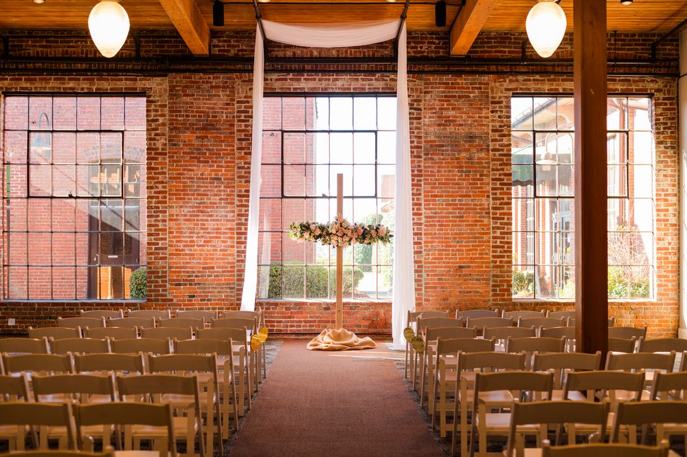 Rustic wedding ceremony venue with exposed brick walls, wooden chairs, and a floral cross altar near large windows.