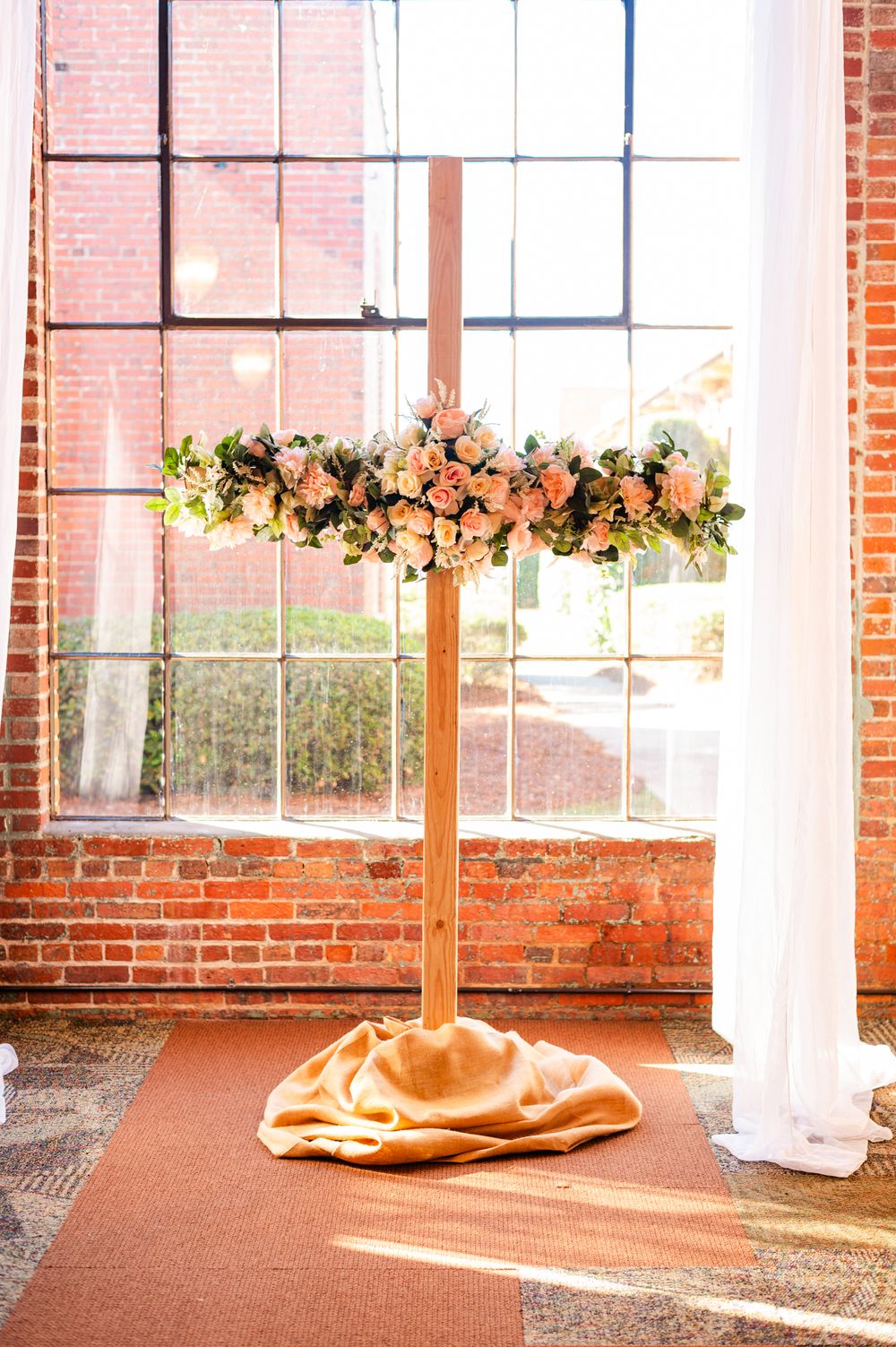 Elegant floral cross centerpiece with pink roses and greenery on a gold base, set against a brick wall backdrop.