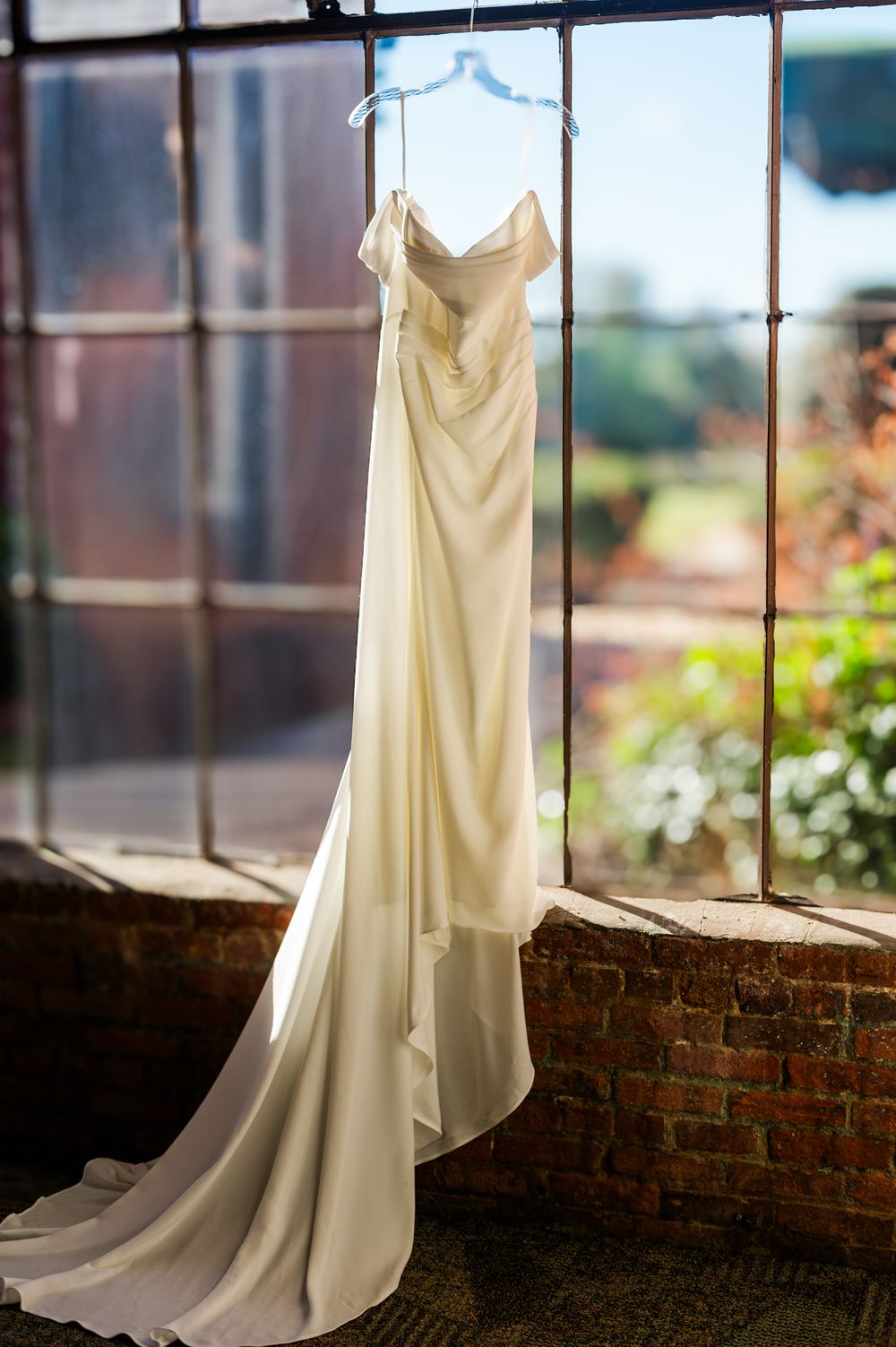 Elegant ivory satin wedding gown with a long train hanging in a sunlit window with brick walls and garden views.