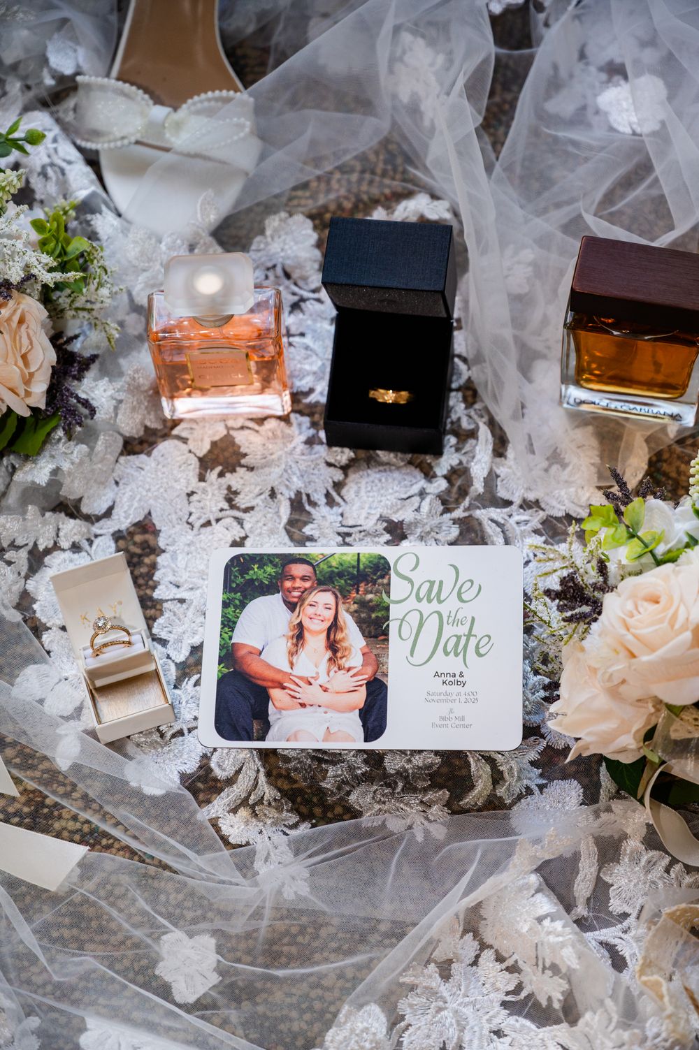 Save the date card with couple photo surrounded by wedding rings, perfume bottles, and white floral veil on lace fabric.