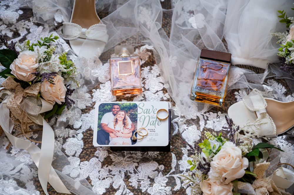 Wedding flat lay with bridal bouquet, veil, photo card, rings, and accessories on white floral backdrop.