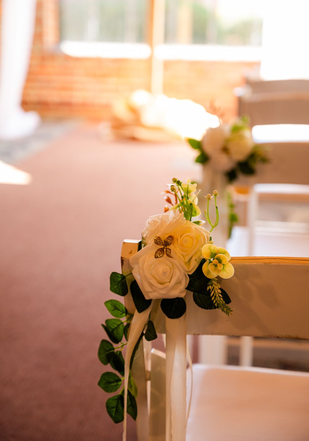 White roses and greenery floral arrangement decorating white wedding ceremony chairs along the aisle.