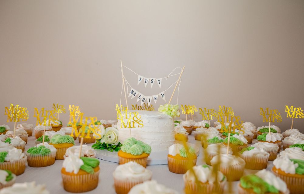 Wedding cake with Mr. & Mrs. topper surrounded by decorated green and yellow frosted cupcakes on a white table.