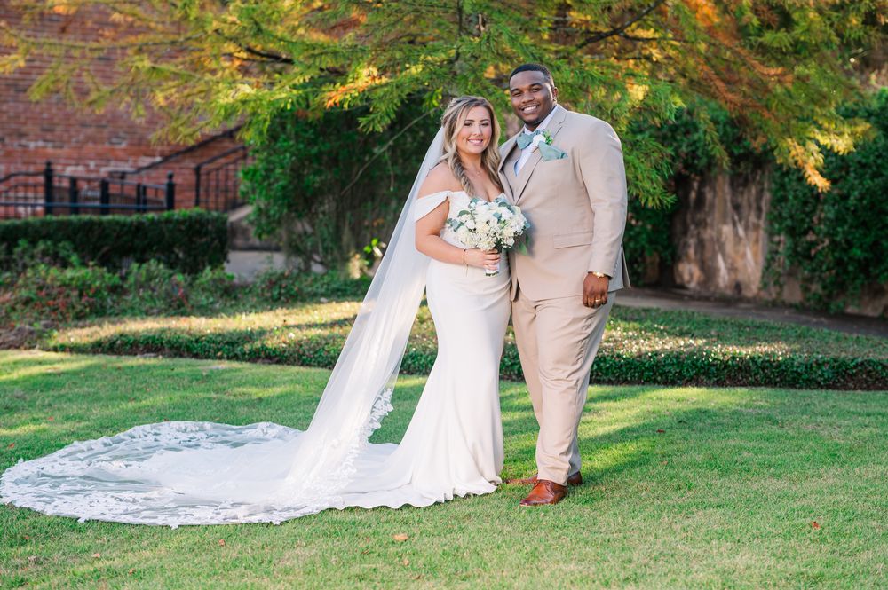 Smiling bride holding white bouquet and groom in tan suit stand together on grass with autumn trees behind them.