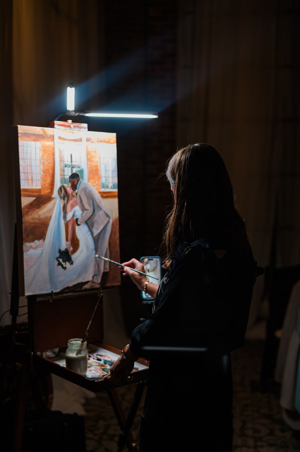 Artist painting a live wedding portrait on canvas at an easel during a dimly lit wedding reception.