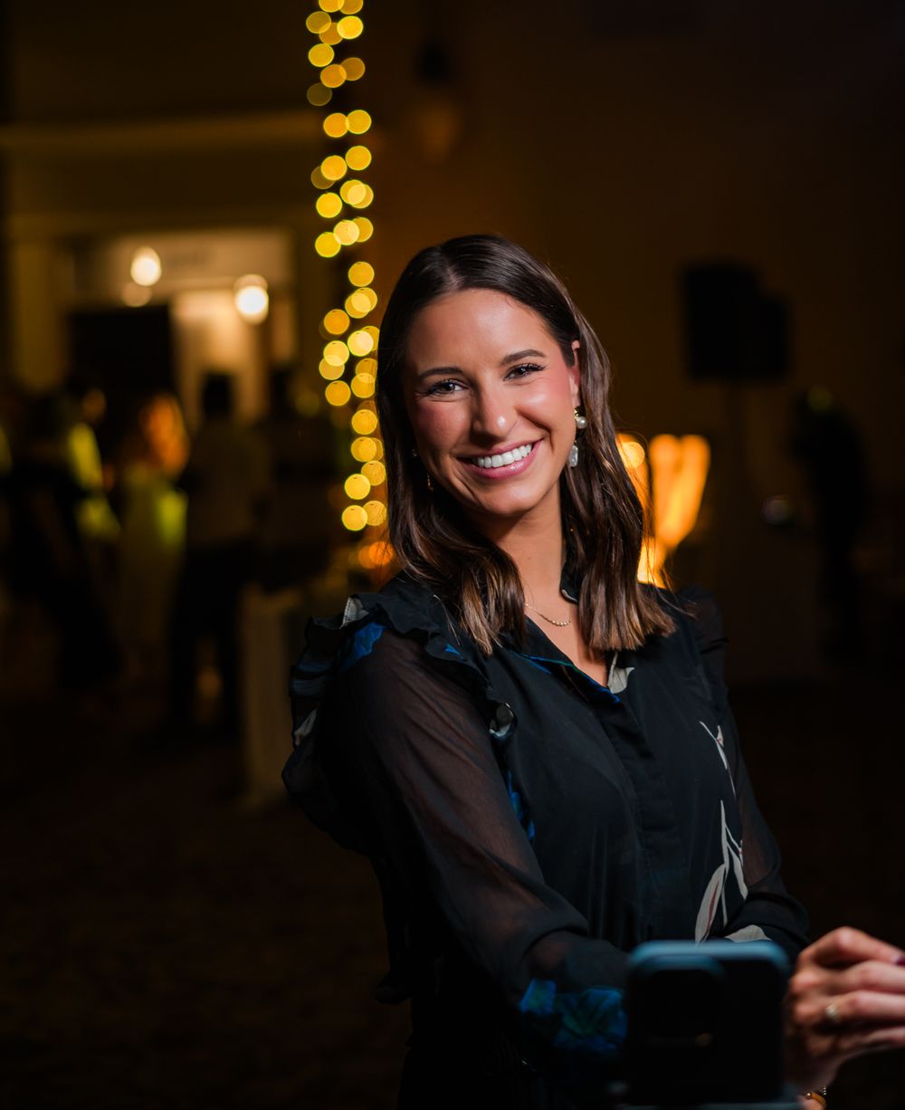 Smiling brunette woman in black outfit at elegant evening event with bokeh string lights background.