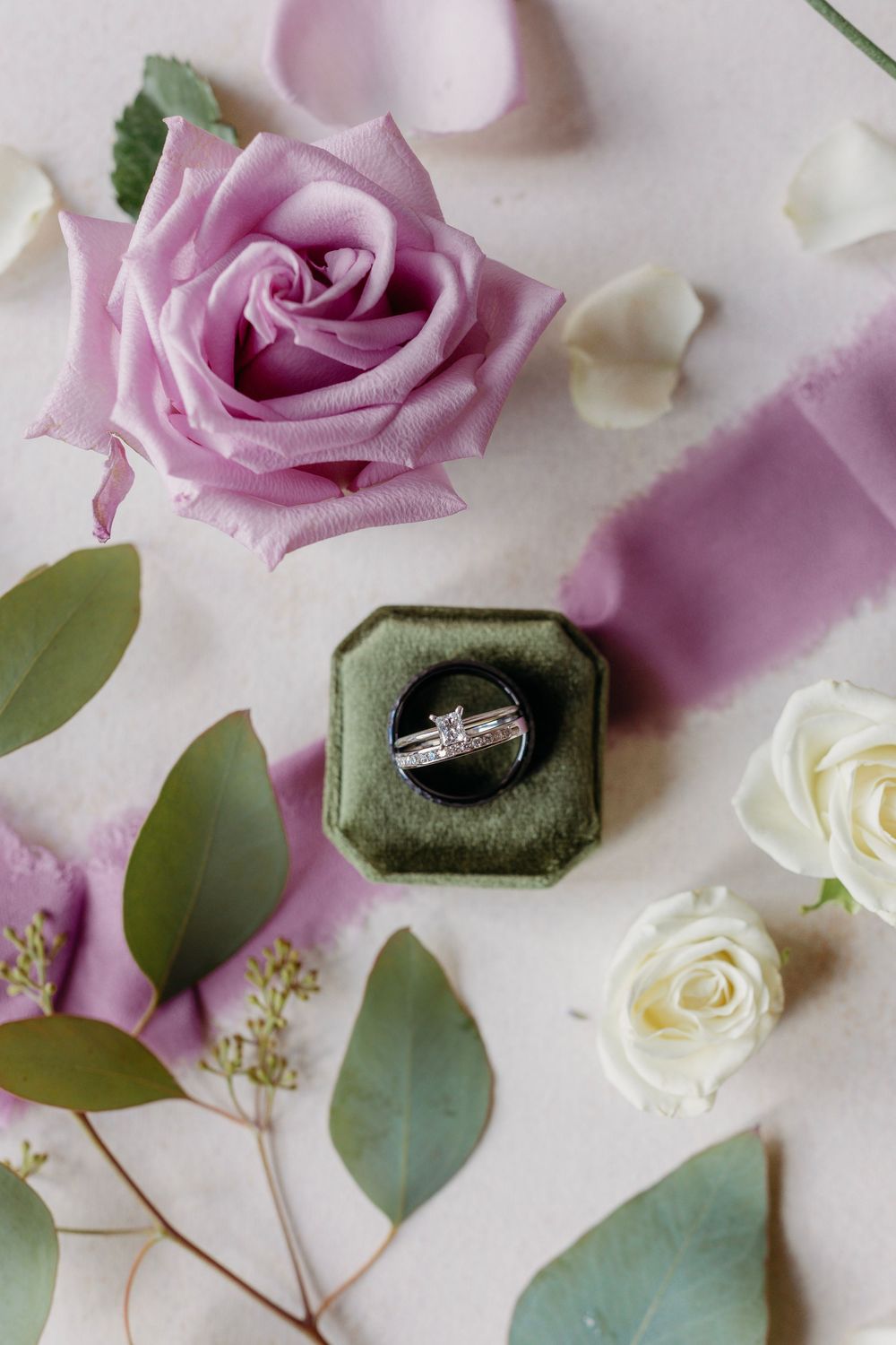 Diamond engagement ring in green velvet box with purple and white roses, eucalyptus leaves, and pink ribbon on white surface.