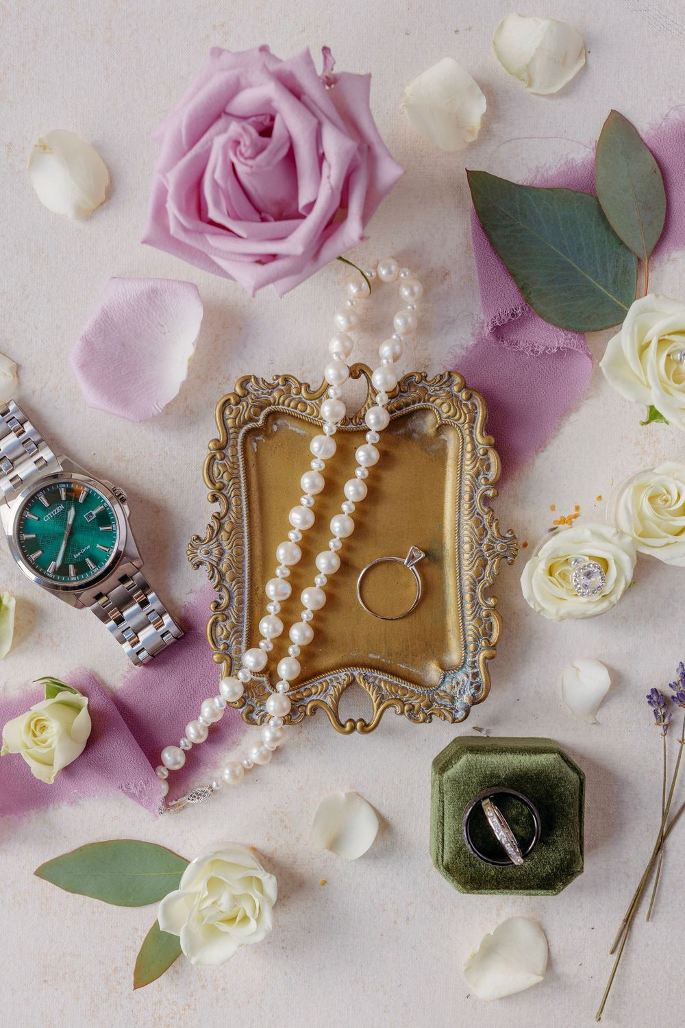 Elegant wedding flat lay with pearl necklace, diamond ring, watch, and white and lavender roses on a soft pink background.