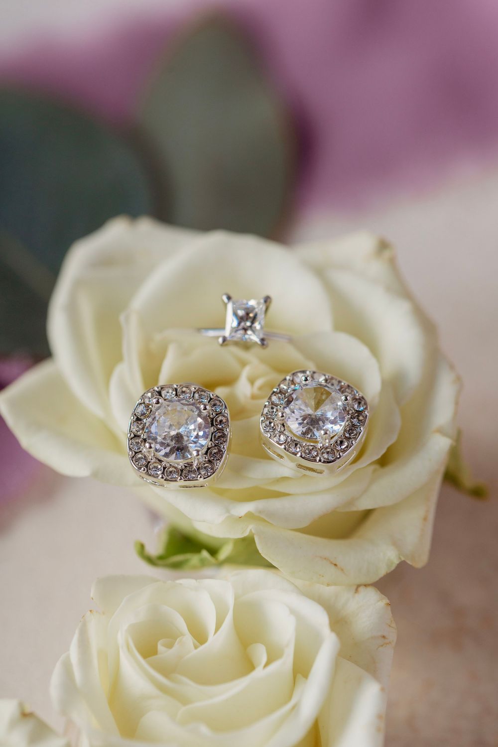 Diamond halo stud earrings and solitaire ring displayed on white roses for a wedding.