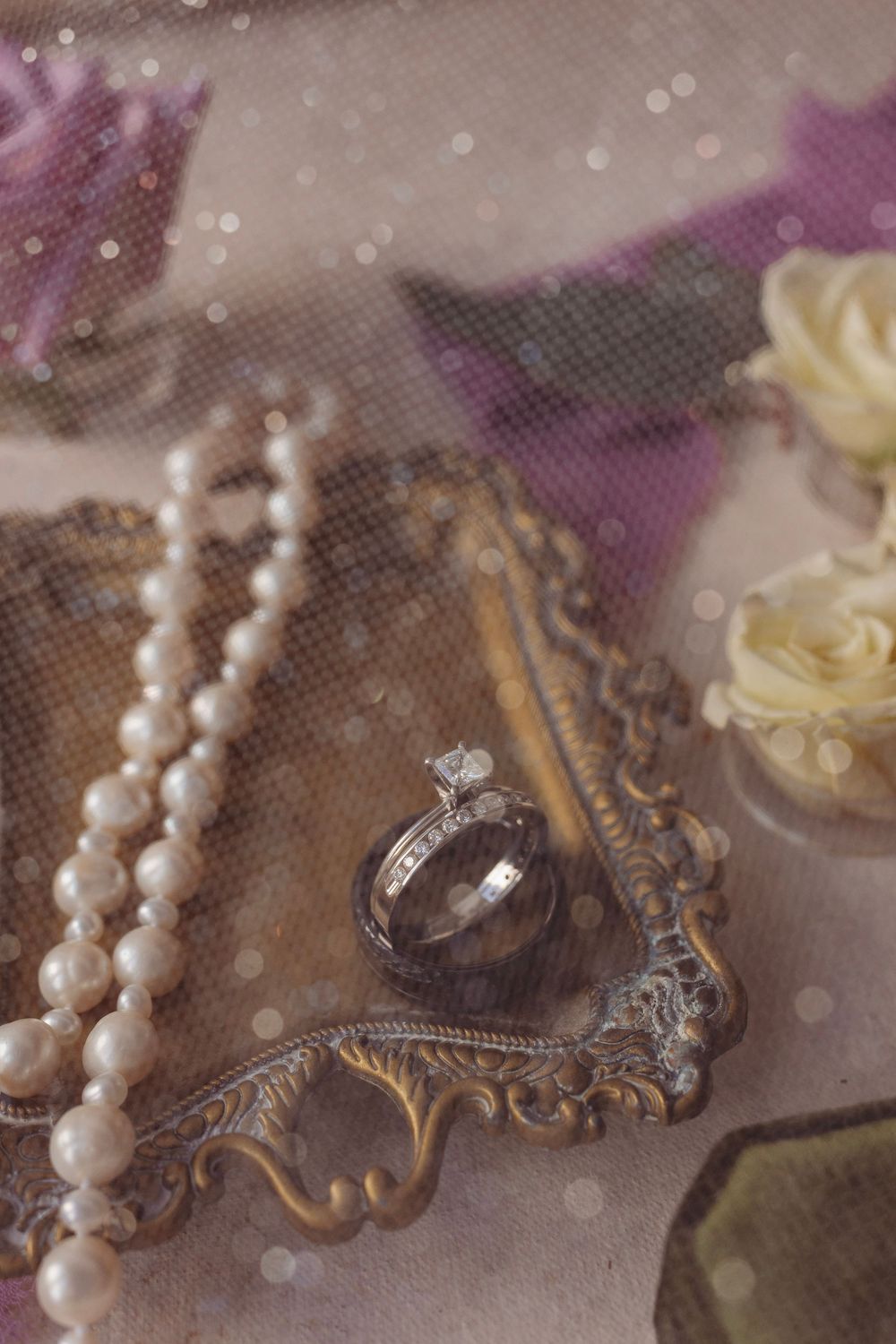 Vintage wedding flat lay with diamond engagement ring, pearl necklace, and white rose on ornate gold mirror tray.