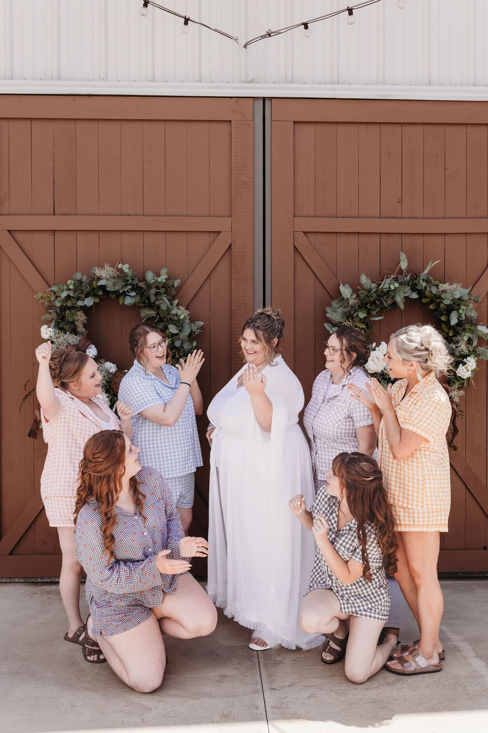 Bride in white gown surrounded by bridesmaids in colorful gingham outfits outside rustic barn doors with floral wreaths.