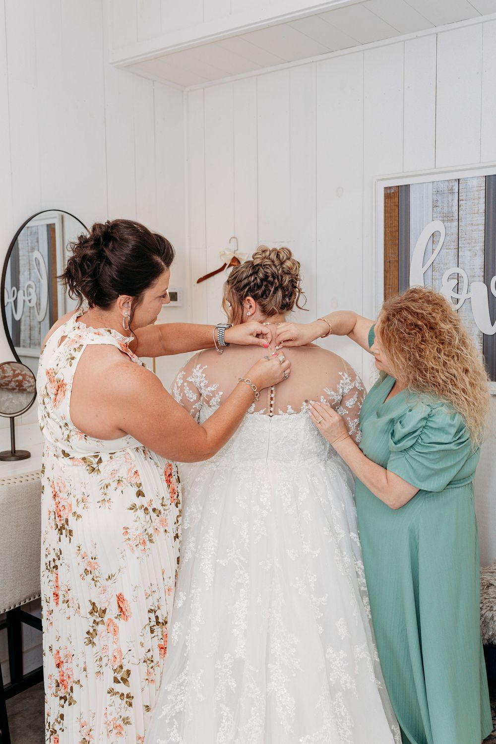 Two women helping a bride button up her lace wedding dress in a bright white bridal suite.