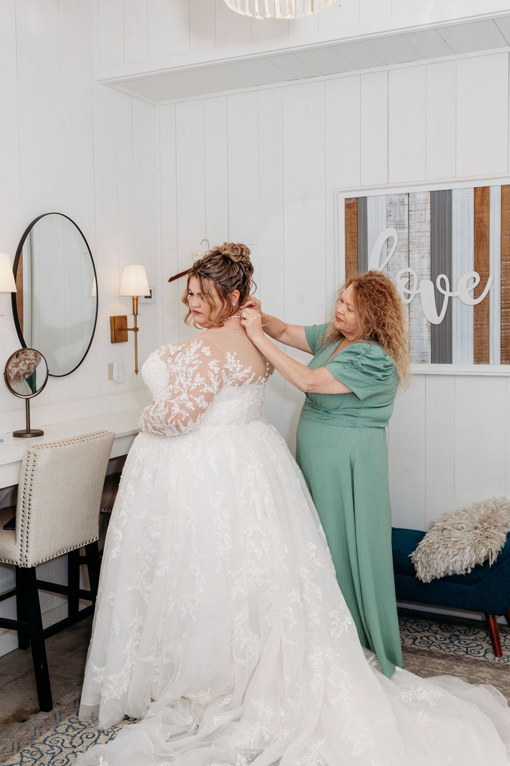 Bride in lace wedding gown gets help with dress from woman in green at a bridal suite with love sign decor.