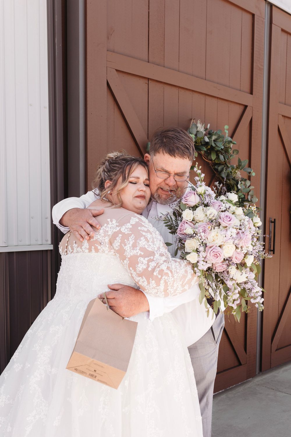 Emotional father-daughter first look embrace outside barn doors, bride holding lavender and white floral bouquet.