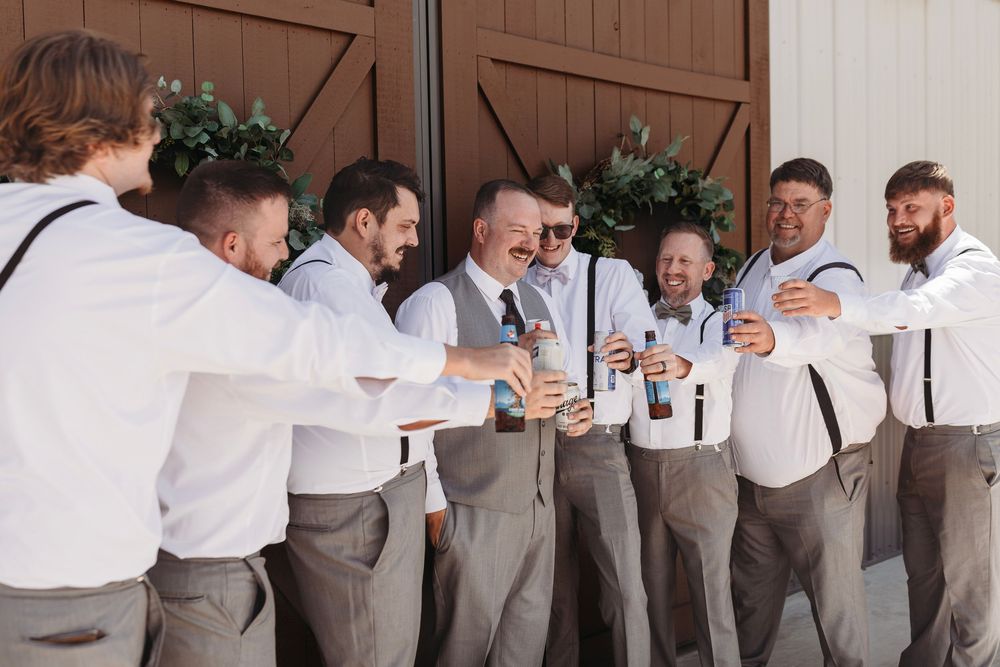 Groomsmen in white shirts and suspenders laughing and toasting with drinks outside a rustic barn venue.